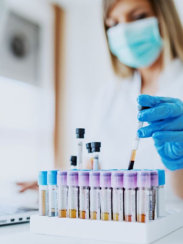 Lab assistant in uniform, wearing a mask and rubber gloves, seated with a blood-filled test tube, typing on a laptop. Test tubes in foreground, selectively focused.